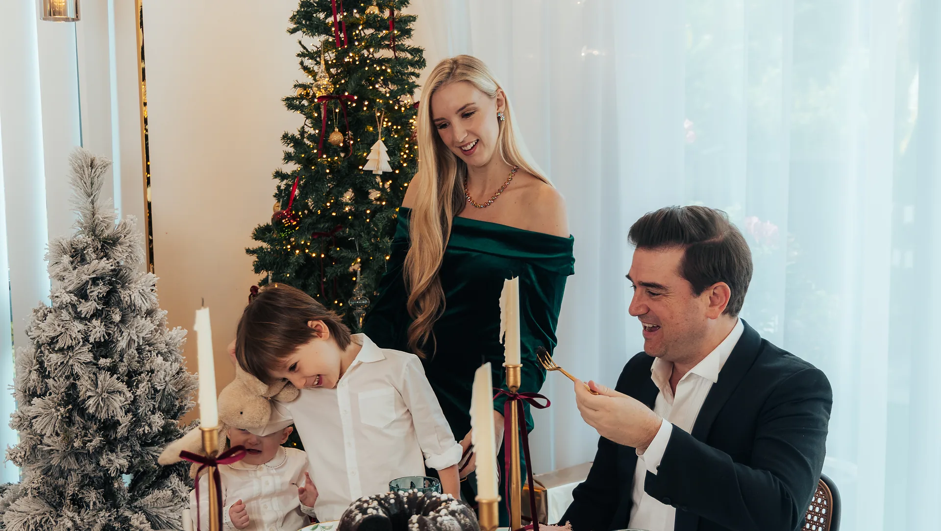 A family of four laughing around a Christmas table, a lit tree behind them