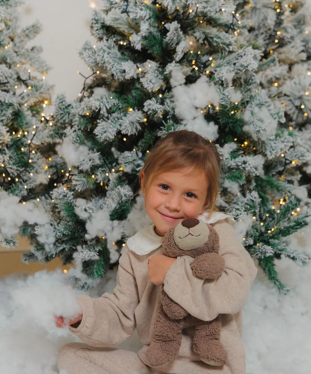 A small girl in cream sitting in deep snow with a teddy, lit flocked trees behind her