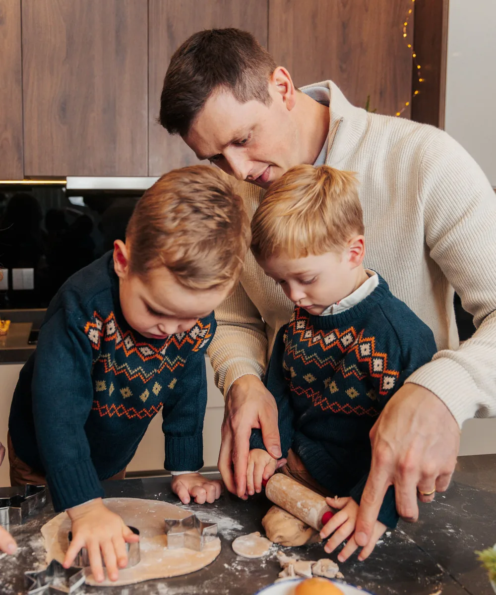 A father and two boys in fair isle jumpers cutting gingerbread on a floured counter