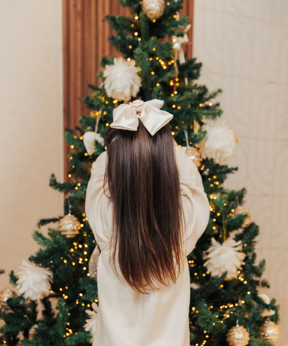 A girl in a cream dress reaching up to hang an ornament on the tree